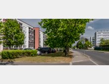 Straßenszene mit mehrstöckigen Wohngebäuden auf der linken Seite, einem Gehweg, geparkten Autos und grünen Bäumen entlang der Straße. Im Hintergrund sind weitere Wohnhäuser und eine klare, teilweise bewölkte Himmel zu sehen.