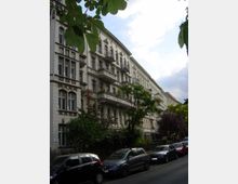 Mehrstöckiges historisches Wohngebäude mit verzierten Fassaden, Balkonen und hohen Fenstern in einer städtischen Straße. Im Vordergrund stehen geparkte Autos, und Bäume säumen den Gehweg.