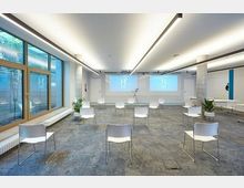 Moderner Seminarraum mit weiß gestrichenen Wänden, Deckenleuchten und großen Fenstern auf der linken Seite, die Tageslicht hereinlassen. Stühle sind mit Abstand angeordnet, und vorne sind zwei Projektionsflächen sowie ein Rednerpult sichtbar.