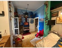 Wohnzimmer mit bunter, individueller Gestaltung. Eine Wand ist in Blau und Grün gestrichen, mit gemalter Meereswellen-Kunst und hängenden Holzkunstwerken dekoriert. Im Raum befinden sich ein roter Sessel, ein kleines Sofa mit Decken, ein Wandfernseher, eine geöffnete weiße Tür, die in einen weiteren Raum führt, sowie Möbel wie ein kleiner Tisch und Regale.