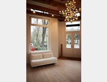 Modernes Wohnzimmer mit hellem Holzboden, weißem Sofa, großen Fensterflächen und einer Glastür, die den Blick auf eine Außenumgebung mit Bäumen und parkenden Autos freigibt. Eine auffällige, bunte Hängelampe ziert die Decke mit Holzbalken.