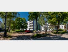 Wohnsiedlung mit mehrstöckigen, hellen Mehrfamilienhäusern und Balkonen, umgeben von hohen Laubbäumen. Im Vordergrund liegt ein kleiner Parkplatz mit mehreren Autos und eine asphaltierte Zufahrt zwischen Grünflächen.