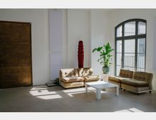 Heller, minimalistischer Lounge- oder Aufenthaltsraum mit weißen Wänden, zwei beigefarbenen Sofas und einem niedrigen weißen Couchtisch auf glattem Betonboden. Ein großes bogenförmiges Fenster mit schwarzem Rahmen lässt viel Tageslicht herein; daneben stehen Topfpflanzen und schlanke Wandpaneele, links eine große braune Tür.