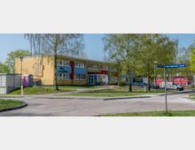 Flaches zweigeschossiges Gebäude mit gelb- und blaufarbiger Fassade, umgeben von Rasenflächen und Bäumen; im Vordergrund eine Straßenecke mit Straßenschildern und Gehwegen.