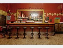 Barbereich in einem nostalgischen, rot gestrichenen Raum mit goldenen Verzierungen. Vor der Theke stehen eine Reihe schwarzer Barhocker, dahinter ein großer Spiegel eingerahmt mit einem goldenen Rahmen, der diverse Flaschen, Gläser und Dekoration reflektiert.