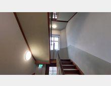 Ein Treppenhaus mit weißen Wänden und grünlicher Wandfarbe im unteren Bereich. Die Treppe hat ein Holzgeländer, ein rechteckiges Fenster beleuchtet den Bereich, und ein Notausgangsschild ist sichtbar.