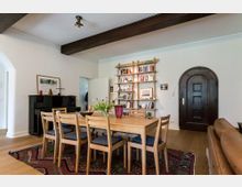 Esszimmer in einem Wohnraum mit einem Holztisch und sechs Stühlen auf einem gemusterten Teppich. Im Hintergrund ein dunkles Klavier, ein offenes Regal mit Büchern und Dekoration sowie eine dunkelbraune, gewölbte Holztür.