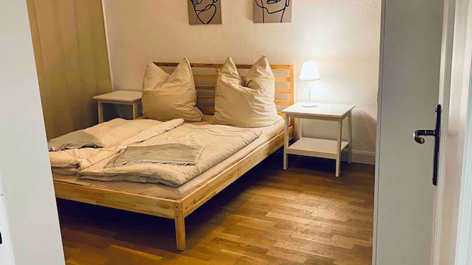 Schlichtes Schlafzimmer mit Holzbett, weißen Kissen und Bettwäsche auf dunklem Holzboden. An der weißen Wand hängen zwei abstrakte Bilder; daneben stehen zwei kleine Nachttische mit Lampe, und rechts ist ein hoher Kleiderschrank sowie eine offene Tür zu sehen.