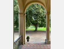 Blick von einer offenen Arkade mit Rundbögen und Säulen auf einen Baum im Grünen, mit einem sandigen Vorplatz, einem kleinen runden Brunnen im Hintergrund und einem dekorativen Pflanzkübel in der Nähe. Der Boden der Arkade ist mit rot-beigen Kacheln bedeckt.