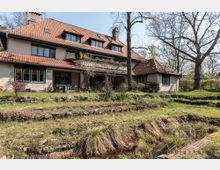 Großes, freistehendes Haus mit rotem Ziegeldach und mehreren Gauben, umgeben von einem weitläufigen Garten mit Rasenflächen, Beeten und Terrassen. Im Vordergrund sind Steinstufen und ein kleiner Wassergraben sichtbar, im Hintergrund stehen blattlose Bäume.