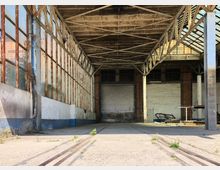 Verlassene Lagerhalle mit rostigen Stahlträgern, großen Glasfenstern und einer geschlossenen Metallrolltür im Hintergrund. Der Betonboden ist teilweise von Pflanzen bewachsen, und die Decke zeigt freiliegende Stahlverstrebungen.