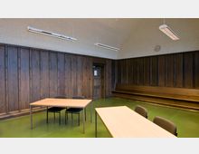 Ein einfach eingerichteter Raum mit Holzwänden, grünem Boden und einer Holzbank entlang der Wand. Zwei Tische und mehrere Stühle stehen im Vordergrund, während an der Decke Hängelampen angebracht sind und eine Wanduhr sichtbar ist.