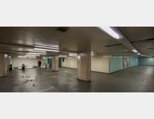 Unterirdischer U-Bahn-Gang mit dunklem Fliesenboden, gelblichen Wänden und säulenartiger Struktur. Neonlampen beleuchten den Raum, eine Rolltreppe befindet sich links, während auf dem Boden verstreute Blätter zu sehen sind.