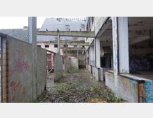 Eine verlassene, heruntergekommene Industriehalle mit teilweise eingestürztem Dach, beschädigten Wänden und Graffiti. Der Boden ist mit Laub und Pflanzen bewachsen, und Metallträger ragen aus der Struktur hervor.