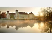 Schloss an einem ruhigen See im Morgennebel, mit hellen Fassaden, Türmen und roten Dächern, umgeben von Bäumen in Herbstfarben. Die reflektierte Silhouette des Gebäudes ist im Wasser zu sehen, während die Sonne durch die Bäume scheint.