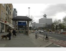 U-Bahnhof-Eingang „Theodor-Heuss-Platz“ mit blauem Schild und umliegender Straßenkreuzung im Vordergrund; links sind Gebäude mit Geschäften, rechts eine mehrspurige Straße mit Autos sowie das markante Hochhaus des Rundfunks Berlin-Brandenburg (rbb).