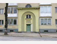 Fassadenansicht eines mehrstöckigen Wohnhauses mit gelber Putzfassade, rechteckigen weißen Fenstern und einem grünen Hauseingang. Der Eingang ist durch einen großen Rundbogen und kleinere Fenster hervorgehoben, während Graffiti und ein Baumstamm im Vordergrund sichtbar sind.