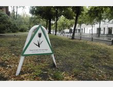 Ein kleiner Park mit Rasenflächen und Bäumen, im Vordergrund ein dreieckiges Schild mit der Aufschrift „Geschützte Grünanlage – Gesetz vom 24.11.1997“ und einer stilisierten Tulpe. Im Hintergrund sind Gehwege, ein Gebäude mit weißer Fassade und ein geparktes Auto zu sehen.