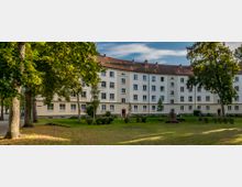 Wohnhaus im Stil des sozialen Wohnungsbaus, mit heller Fassade und roten Ziegeldächern, in einer Grünanlage mit Rasen, Bäumen und einem kleinen Denkmal im Vordergrund. Der Himmel ist teils bewölkt, und die Umgebung wirkt ruhig und gepflegt.