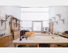 Eine helle Werkstatt mit Holzarbeitstischen, Werkzeugen und aufgestapelten Holzplatten. An den Wänden befinden sich Halterungen und Werkzeuge, während eine gläserne Tür am Ende des Raums Tageslicht hereinlässt.