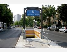 Eingang zur U-Bahn-Station "Konstanzer Straße" mit einem blauen Stationsschild, Treppenabgang und gelb-weiß gestreiften Wandfliesen. Im Hintergrund sind eine Straße mit geparkten Autos, Bäume und ein Hochhaus zu sehen.