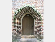 Spitzbogiges Portal aus Backstein einer historischen Kirche oder eines Gebäudes, mit einer Holztür und einem dekorativen Blumenkranz, der den Bogen einrahmt. Die Fassade besteht aus roten Backsteinen, typischer für mittelalterliche Architektur.