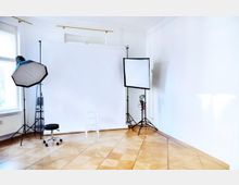 Heller Raum mit Holzparkettboden, eingerichtet als Fotostudio mit einem weißen Hintergrund, einer kleinen weißen Leiter, einem Drehhocker und zwei Fotoleuchten auf Stativen (eine rechteckige und eine achteckige Softbox). Links befindet sich ein großes Fenster.