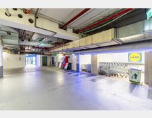 Eingangsbereich eines Einkaufszentrums oder Supermarkts mit Betonboden und sichtbaren Rohren an der Decke. Rechts sind Einkaufswagen unter einer beleuchteten Markierung mit dem Logo eines Lidl-Marktes sichtbar, links führt ein Ausgang ins Freie.