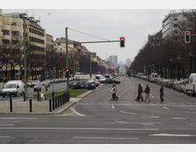 Weitläufige städtische Straße mit mehrspurigen Fahrbahnen und parkenden Autos entlang beider Seiten, flankiert von mehrstöckigen Gebäuden und kahlen Bäumen; im Hintergrund ist der Berliner Fernsehturm sichtbar. Fußgänger überqueren einen Zebrastreifen bei einer roten Ampel im Vordergrund.