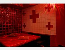Ein rot beleuchteter Raum mit einer roten Liege, darauf zwei Kissen. Die Wand rechts ist mit weißen Kacheln verkleidet, die rote Kreuze formen, während die linke Wand mit Glasbausteinen gestaltet ist.