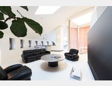 Moderner Wohnraum mit hellen Wänden und großen Oberlichtern, eingerichtet mit schwarzen Ledersesseln, einem runden Metalltisch und einer offenen Küche im Hintergrund. Links stehen kleine Fenster in einer weißen Wandreihe, während der Raum rechts durch rosafarbene Vorhänge und Säulen begrenzt wird.