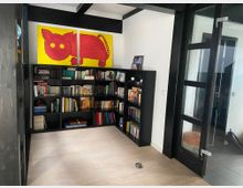 Ein kleiner Raum mit hellen Holzböden, einer schwarzen Bücherwand voller Bücher und einem auffälligen, gelben Gemälde mit einer roten Katze an der Wand. Rechts befindet sich eine verglaste Tür mit schwarzem Rahmen.