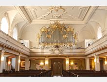 Innenraum einer historischen Kirche mit Blick auf eine große, goldverzierte Orgel auf der Empore. Die Decke ist mit weißen Stuckelementen verziert, und der Raum wird von großen Rundbogenfenstern sowie hölzernen Kirchenbänken geprägt.