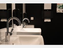 Ein öffentliches Waschbecken mit glänzenden Metallarmaturen und weißer Keramik vor einer schwarzen Wand. Im Hintergrund sind Seifenspender, ein Papierhandtuchspender und ein Spiegel zu sehen.