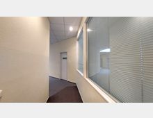 Ein schmaler Büroflur mit beigem Anstrich und grauem Teppichboden; rechts verläuft eine Glaswand mit Jalousien, links ist eine geschlossene Wand. Im Hintergrund führt die Tür zu einem weiteren Raum.