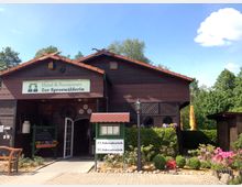 Holzgebäude eines Hotels und Restaurants mit der Aufschrift "Zur Spreewälderin", umgeben von gepflegten Blumenbeeten und Grünflächen; im Vordergrund steht eine Laterne, und im Hintergrund sind Bäume und blauer Himmel zu sehen. Ein Fahrradverleih ist durch Schilder ebenfalls ausgewiesen.