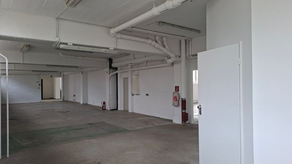 Großer, leerer Innenraum in einem Industriegebäude mit weiß gestrichenen Wänden und Decke, sichtbaren Rohrleitungen und Leuchtstofflampen. Betonboden, mehrere Durchgänge und Türen sowie ein Feuerlöscher an der Wand sind zu sehen.