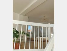 Ein lichtdurchfluteter Dachraum mit weiß gestrichenen Wänden und Holzfußboden. Im Vordergrund eine weiße Treppenbrüstung, dahinter befinden sich ein großes grünes Zimmerpflanze, ein Holzbett und ein kleiner Tisch, mit einem Fenster und einem Dachfenster im Hintergrund.