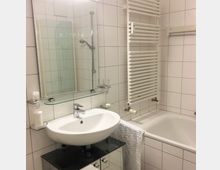 Modernes, weiß gefliestes Badezimmer mit Waschbecken und Wandspiegel samt Glasablage sowie einer Badewanne mit Duscharmatur. An der Wand neben der Wanne befindet sich ein großer, vertikaler Handtuchheizkörper.