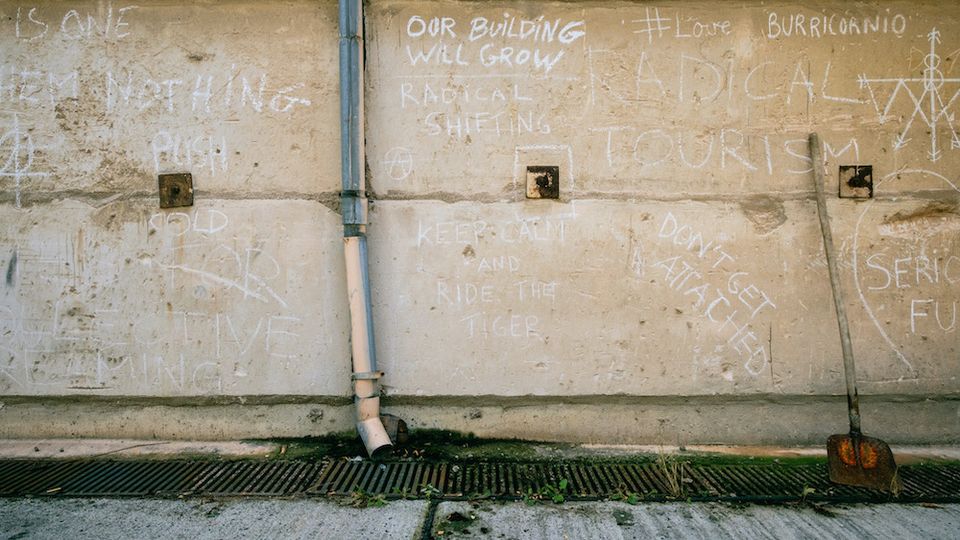 Eine verwitterte Betonwand mit Kreidezeichnungen und -schriftzügen, umgeben von mehreren kleinen quadratischen Wandöffnungen. Ein Fallrohr läuft senkrecht entlang der Wand, und am Boden befinden sich ein Metallgitter mit Abfluss sowie eine Schaufel, die an der Wand lehnt.