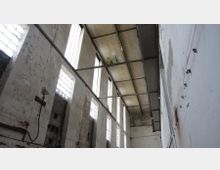 Industriehalle mit hohen, weißen Wänden, die Abnutzungsspuren aufweisen. Große Fenster aus Glasbausteinen und eine Decke mit Stahlträgern lassen Licht herein, während Kabel und Rohre an den Wänden sichtbar sind.