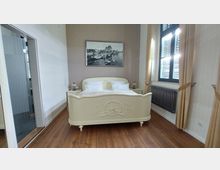 Schlafzimmer mit einem cremefarbenen Bett im antiken Stil, verziert mit floralen Ornamenten, flankiert von zwei Nachttischen mit Lampen. Im Hintergrund hängt ein Schwarz-Weiß-Bild mit Hafenszene, und rechts befindet sich ein großes Fenster mit beigen Vorhängen und einem Heizkörper darunter.