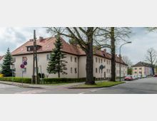 Straßenszene in einem Wohngebiet mit niedrigen, hellen Mehrfamilienhäusern und roten Ziegeldächern; im Vordergrund stehen Bäume, Verkehrszeichen und parkende Autos am Straßenrand. Die Straße führt an einer Kreuzung entlang, gesäumt von Gehwegen und gepflegten Grünflächen.