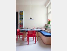 Essbereich in einer Ecke mit einem ovalen Tisch, zwei roten Kunststoffstühlen und einer Holzbank mit blauen Polsterauflagen. Im Hintergrund ein Regal mit Büchern und Dekorationen, eine Hängelampe mit schwarzem Schirm sowie ein Fenster, durch das Tageslicht einfällt; darauf eine Vase mit roten Tulpen.