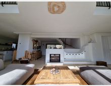 Wohnzimmer in einem modernen Haus mit hellen Wänden und Holzelementen. In der Mitte befindet sich ein Kamin, umgeben von einer geschwungenen weißen Wand, dahinter eine offene Treppe aus Holz und Metall; links ist eine offene Küche zu sehen.