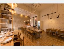 Gemütlicher Essbereich mit langen Holztischen und Stühlen auf einem Holzboden, umgeben von weißen und ziegelroten Backsteinwänden. Die Decke ist teilweise mit Lochplatten gestaltet, und hängende Glühbirnen sorgen für warme Beleuchtung; an der Wand befinden sich Regale mit Geschirr und ein Kühlschrank.