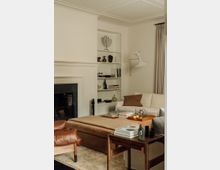 Wohnzimmer mit eleganter Inneneinrichtung: Ein Kamin auf der linken Seite wird von weißen Wänden und einem hellen Einbauregal mit dekorativen Objekten wie Vasen und einer Metall-Menora flankiert. Ein cremefarbenes Sofa, ein brauner Polsterhocker, ein Ledersessel und ein Beistelltisch mit Büchern und Skulpturen prägen den Raum, der dezent beleuchtet ist.