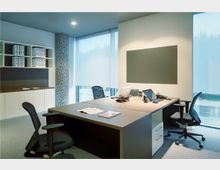 Modernes Büro mit einem großen, hölzernen Schreibtisch, zwei schwarzen Bürostühlen und mehreren Computern und Telefonen. Im Hintergrund befinden sich ein Regal mit Ordnern, ein Mosaiksäulen-Detail und große Fenster mit halbtransparenten Rollos.