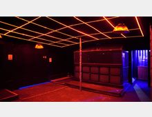 Ein dunkler Innenraum, vermutlich einer Bar oder eines Clubs, mit einer Decke aus leuchtenden roten Linien in einem Gittermuster. Im Vordergrund eine erhöhte Plattform mit Metallgittern, dahinter ein beleuchteter Bereich in Blau und eine Theke.