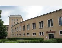 Gebäude im Stil der klassischen Moderne mit heller Steinfassade und rechteckigen Fenstern. Im Vordergrund ist ein Eingang mit der Aufschrift „Silver Wings“ sichtbar, umgeben von gepflegten Grünanlagen.